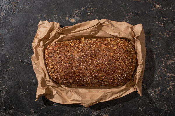 Vista superior de la hogaza recién horneada de pan de grano entero en papel sobre la superficie de piedra negro - foto de stock