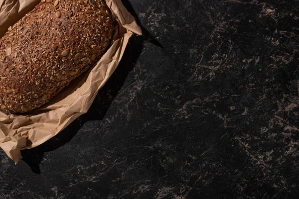 Top view of fresh baked loaf of whole grain bread in paper on stone black surface — Stock Photo