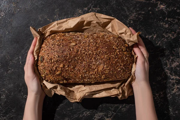 Vista cortada da mulher segurando pão cozido fresco do pão de grão inteiro no papel na superfície preta da pedra — Fotografia de Stock