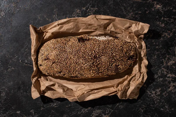 Top view of fresh baked loaf of whole grain bread with sesame in paper on stone black surface — Stock Photo