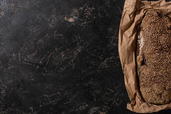 Top view of fresh baked loaf of whole grain bread with sesame in paper on stone black surface — Stock Photo