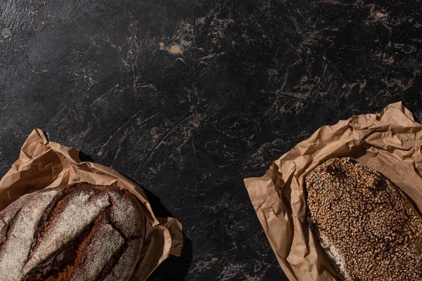 Vista superior de los panes de pan de grano entero recién horneados en papel sobre la superficie negra de piedra - foto de stock