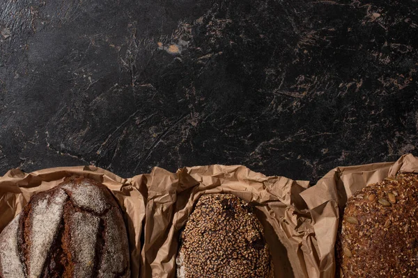 Vue du dessus des pains de pain frais à grains entiers cuits au four en papier sur une surface noire en pierre — Photo de stock