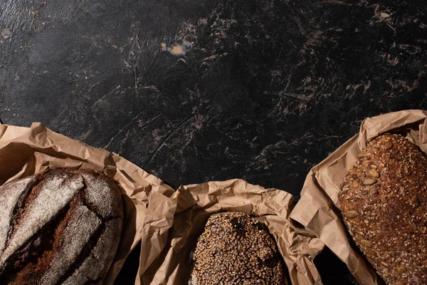 Vue du dessus des pains de pain frais à grains entiers cuits au four en papier sur une surface noire en pierre — Photo de stock