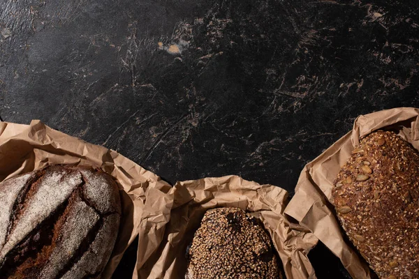 Top view of fresh baked whole grain bread loaves in paper on stone black surface — Stock Photo