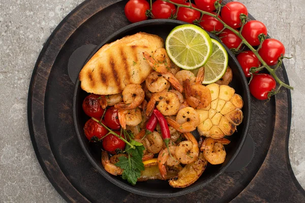 Draufsicht auf gebratene Garnelen mit gegrillten Toasts, Gemüse, Kirschtomaten und Limette auf Holzbrett auf grauem Betongrund — Stockfoto