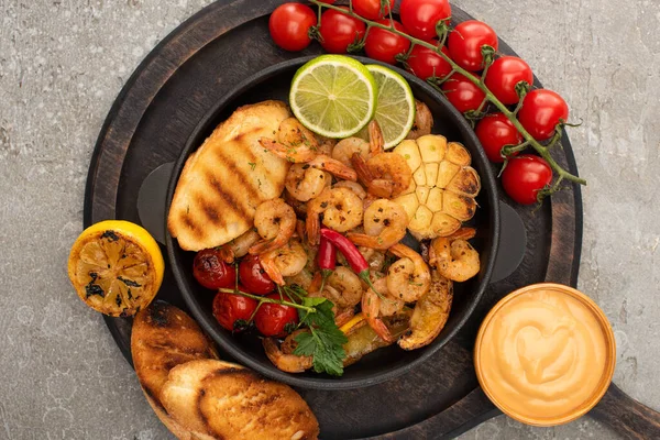 Top view of fried shrimps with grilled toasts, vegetables, cherry tomatoes and lime on wooden board with sauce on grey concrete background — Stock Photo