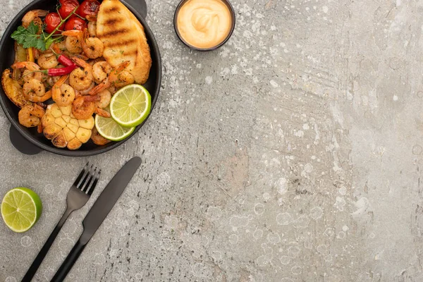 Vista superior de camarones fritos con tostadas a la parrilla, verduras y lima cerca de cubiertos y salsa sobre fondo de hormigón gris - foto de stock