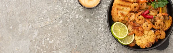 Top view of fried shrimps with grilled toasts, vegetables and lime near sauce on grey concrete background, panoramic shot — Stock Photo