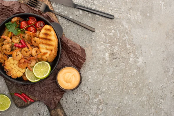 Draufsicht auf gebratene Garnelen mit gegrillten Toasts, Gemüse und Limetten an Bord in der Nähe von Besteck und Sauce auf grauem Betonhintergrund — Stockfoto