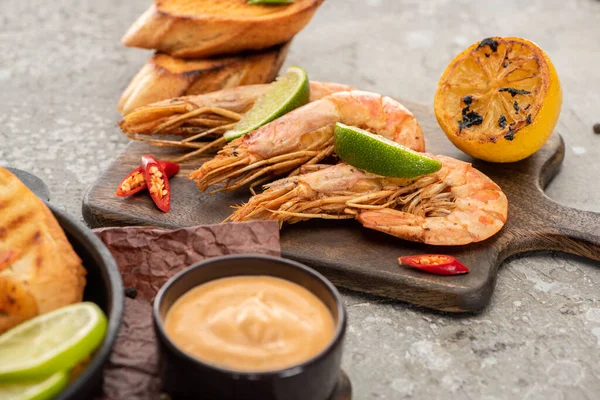 Selective focus of fried shrimps with grilled toasts, lemon and lime on wooden board near sauce on grey concrete background — Stock Photo