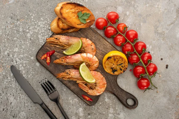 Top view of fried shrimps with grilled toasts, lemon and lime near cherry tomatoes on wooden board near cutlery on grey concrete background — Stock Photo