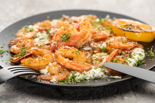 Selective focus of fried shrimps in sauce with dill and lemon in black plate on grey concrete background with cutlery — Stock Photo