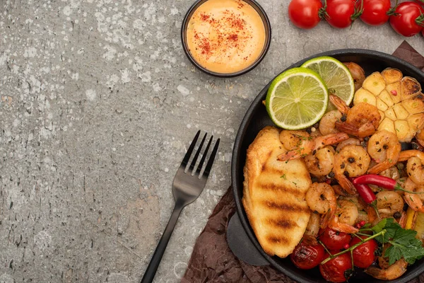 Draufsicht auf gebratene Garnelen mit Sauce, Gabel, gegrillten Toasts, Gemüse und Limette auf grauem Betongrund — Stockfoto