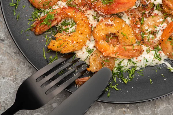 Close up view of fried shrimps in sauce with dill in black plate on grey concrete background with cutlery — Stock Photo