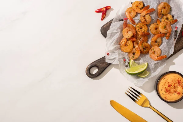 Vue du dessus des crevettes frites sur papier sulfurisé sur carton avec couverts, piment, sauce et citron vert sur fond blanc — Photo de stock