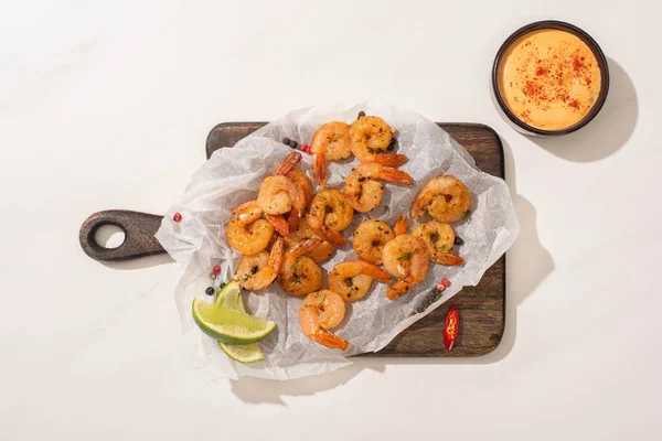 Top view of fried shrimps on parchment paper on wooden board with chili pepper, sauce and lime on white background — Stock Photo