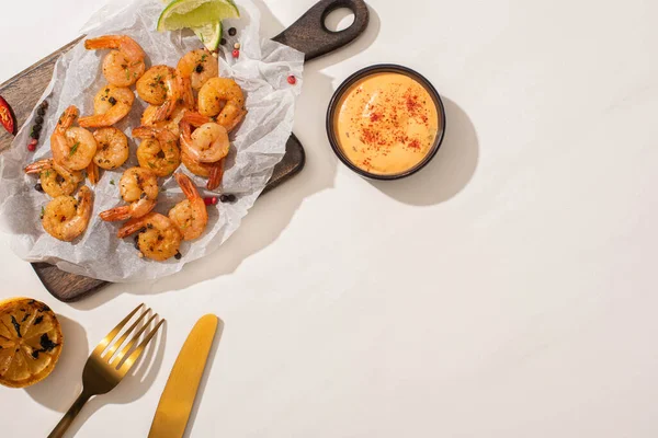 Draufsicht auf gebratene Garnelen auf Pergamentpapier auf Holzbrett in der Nähe gegrillter Zitrone, Sauce und Besteck auf weißem Hintergrund — Stockfoto