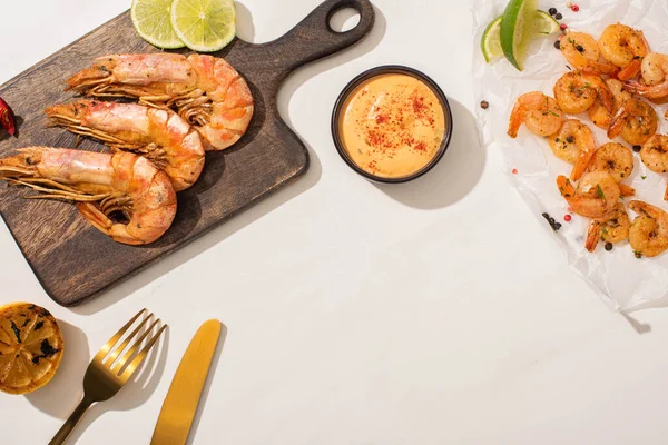 Vue de dessus des crevettes frites sur papier parchemin et carton en bois près du citron grillé, sauce et couverts sur fond blanc — Photo de stock