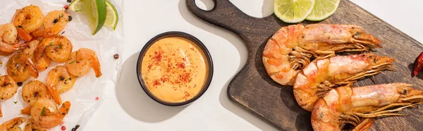 Top view of fried shrimps on parchment paper and wooden board near grilled sauce on white background, panoramic shot — Stock Photo