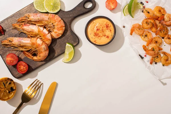 Vue de dessus des crevettes frites sur papier parchemin et carton en bois près du citron grillé, sauce et couverts sur fond blanc — Photo de stock