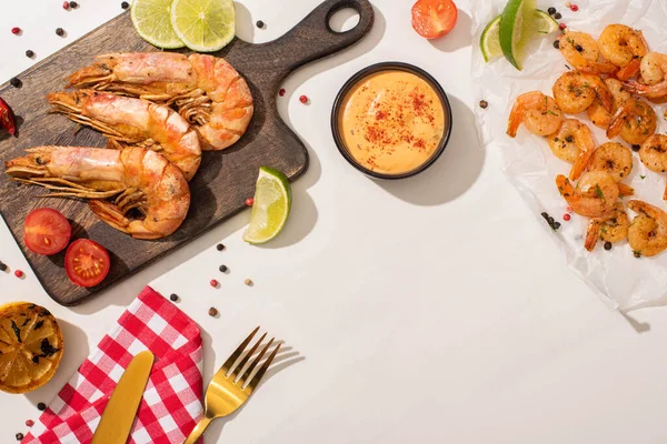 Vista superior de camarões fritos em pergaminho e tábua de madeira com talheres, guardanapo xadrez, legumes, molho e limão no fundo branco — Fotografia de Stock