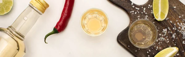 Top view of golden tequila with lime, chili pepper, salt on wooden cutting board on white marble surface, panoramic shot — Stock Photo