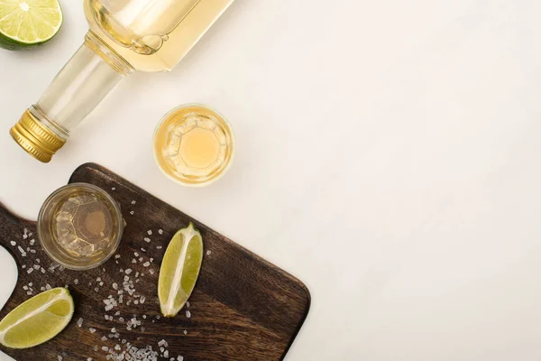 Vista superior da tequila dourada com limão, sal na placa de corte de madeira na superfície de mármore branco — Fotografia de Stock