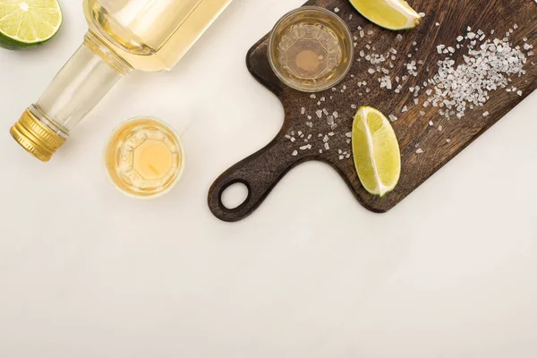 Vista superior da tequila dourada com limão, sal na placa de corte de madeira na superfície de mármore branco — Fotografia de Stock