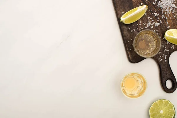 Blick von oben auf goldenen Tequila mit Kalk, Salz auf Holzschneidebrett auf weißem Marmor — Stockfoto