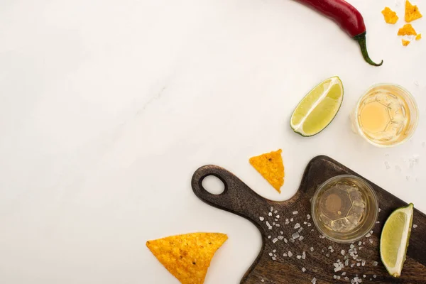 Top view of golden tequila with lime, chili pepper, salt and nachos near wooden cutting board on white marble surface — Stock Photo