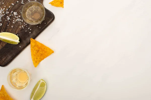 Vista superior da tequila dourada com limão, sal e nachos perto da placa de corte de madeira na superfície de mármore branco — Fotografia de Stock