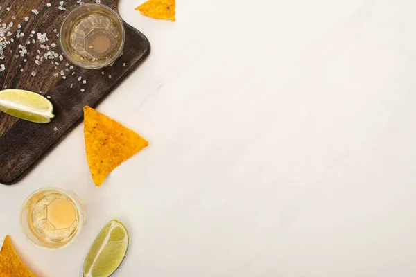 Vista superior da tequila dourada com limão, sal e nachos perto da placa de corte de madeira na superfície de mármore branco — Fotografia de Stock