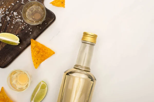 Vista superior da tequila dourada com limão, sal e nachos perto da placa de corte de madeira na superfície de mármore branco — Fotografia de Stock