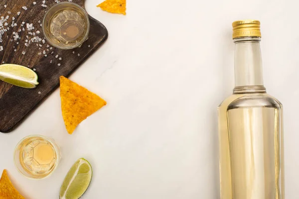 Top view of golden tequila with lime, salt and nachos near wooden cutting board on white marble surface — Stock Photo