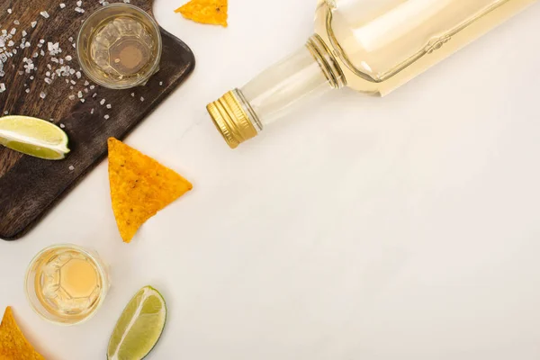 Top view of golden tequila with lime, salt and nachos near wooden cutting board on white marble surface — Stock Photo