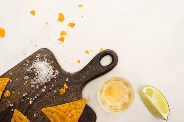 Top view of golden tequila with lime, salt and nachos on wooden cutting board on white marble surface — Stock Photo