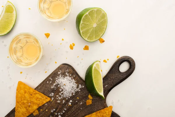Top view of golden tequila in shot glasses with lime, salt and nachos on wooden cutting board on white marble surface — Stock Photo
