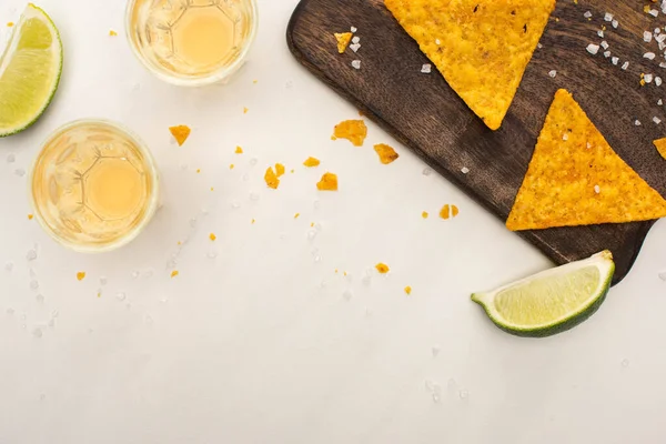 Top view of golden tequila in shot glasses with lime, salt and nachos on wooden cutting board on white marble surface — Stock Photo