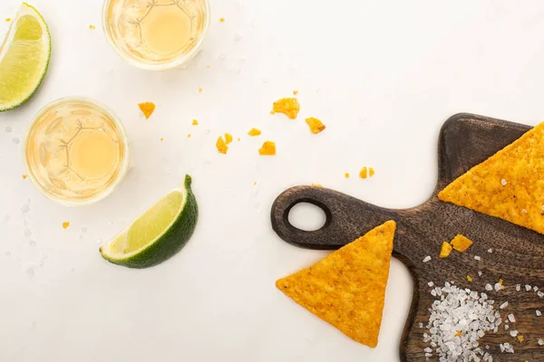 Vue de dessus de la tequila dorée dans des verres de tir avec de la chaux, du sel et des nachos sur une planche à découper en bois sur une surface en marbre blanc — Photo de stock