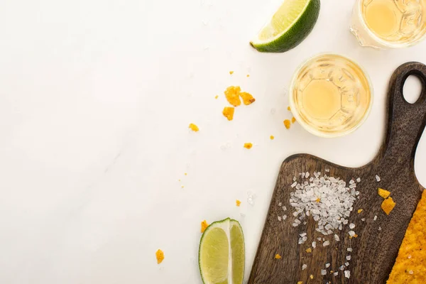 Top view of golden tequila in shot glasses with lime, salt and nachos on wooden cutting board on white marble surface — Stock Photo