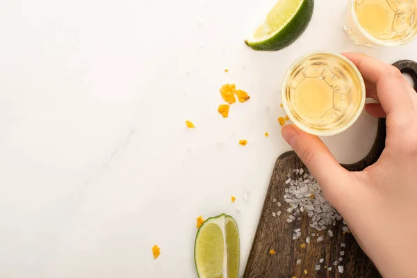 Vista ritagliata di donna che beve tequila dorata con calce, sale sulla superficie di marmo bianco — Foto stock