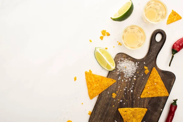 Top view of golden tequila in shot glasses with chili peppers, lime, salt and nachos on wooden cutting board on white marble surface — Stock Photo