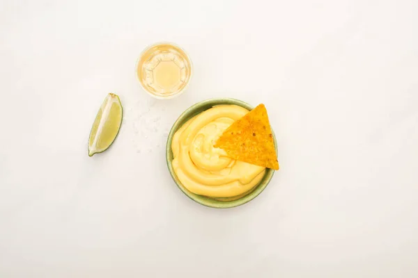 Vista superior de tequila dourada com limão e tigela com molho de queijo e nachos na superfície de mármore branco — Fotografia de Stock