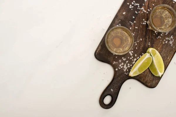 Vista superior da tequila dourada com limão, sal na placa de corte de madeira na superfície de mármore branco — Fotografia de Stock