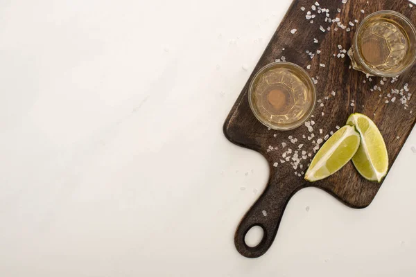 Vue de dessus de tequila dorée à la chaux, sel sur planche à découper en bois sur marbre blanc — Photo de stock