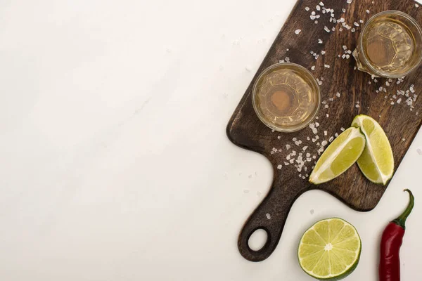 Vista superior de tequila dourada com limão, pimenta, sal na placa de corte de madeira na superfície de mármore branco — Fotografia de Stock