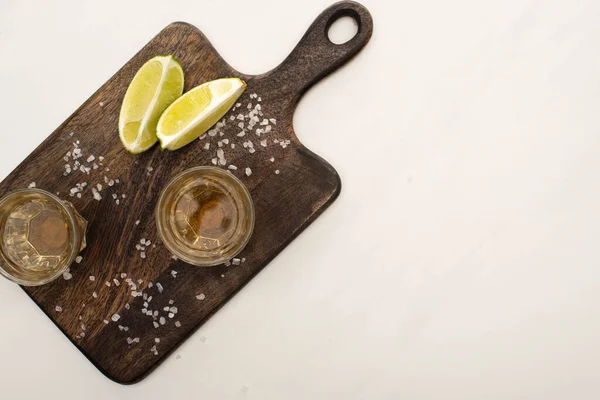 Vue de dessus de tequila dorée à la chaux, sel sur planche à découper en bois sur marbre blanc — Photo de stock