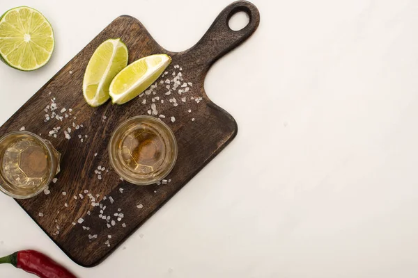 Top view of golden tequila with lime, chili pepper, salt on wooden cutting board on white marble surface — Stock Photo