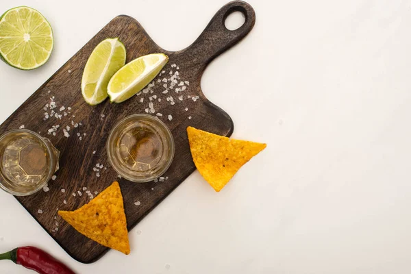 Top view of golden tequila with lime, chili pepper, salt and nachos near wooden cutting board on white marble surface — Stock Photo